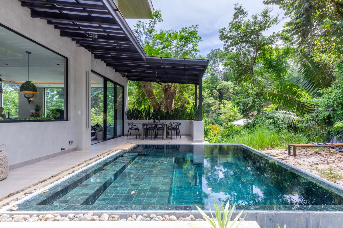 An inviting infinity pool reflects the surrounding greenery, bordered by smooth stones. A covered terrace with dining seating overlooks the pool, enhancing the connection to nature. Floor-to-ceiling windows open to the lush jungle landscape, allowing light to fill the space and create a serene environment.