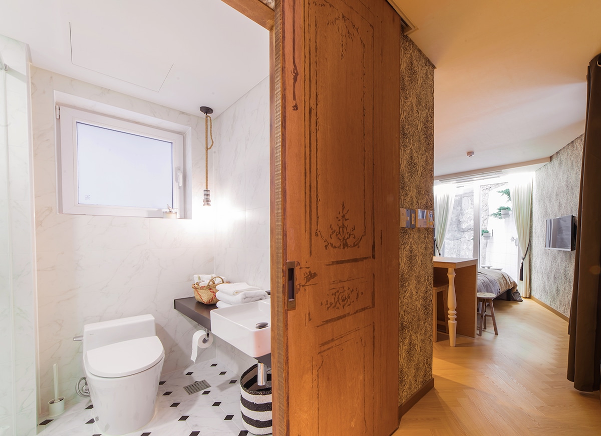 A wooden door opens to a well-designed space featuring a modern bathroom and a cozy sleeping area. The bathroom showcases stylish fixtures on patterned tiles, while soft natural light enters through the window. A dining area is visible, enhancing the inviting layout.