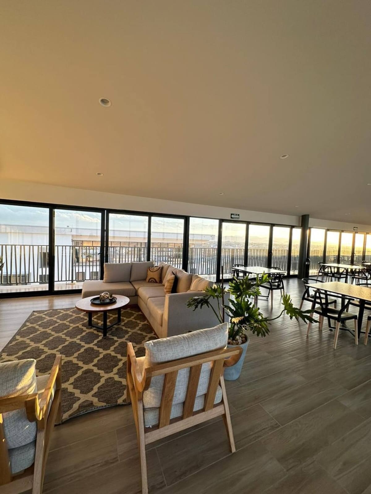 A comfortable lounge area features a sectional sofa and a circular coffee table, complemented by armchairs. Large glass doors provide expansive views and ample natural light. The space is adorned with a patterned rug and potted plants, creating an inviting environment for relaxation or gathering.