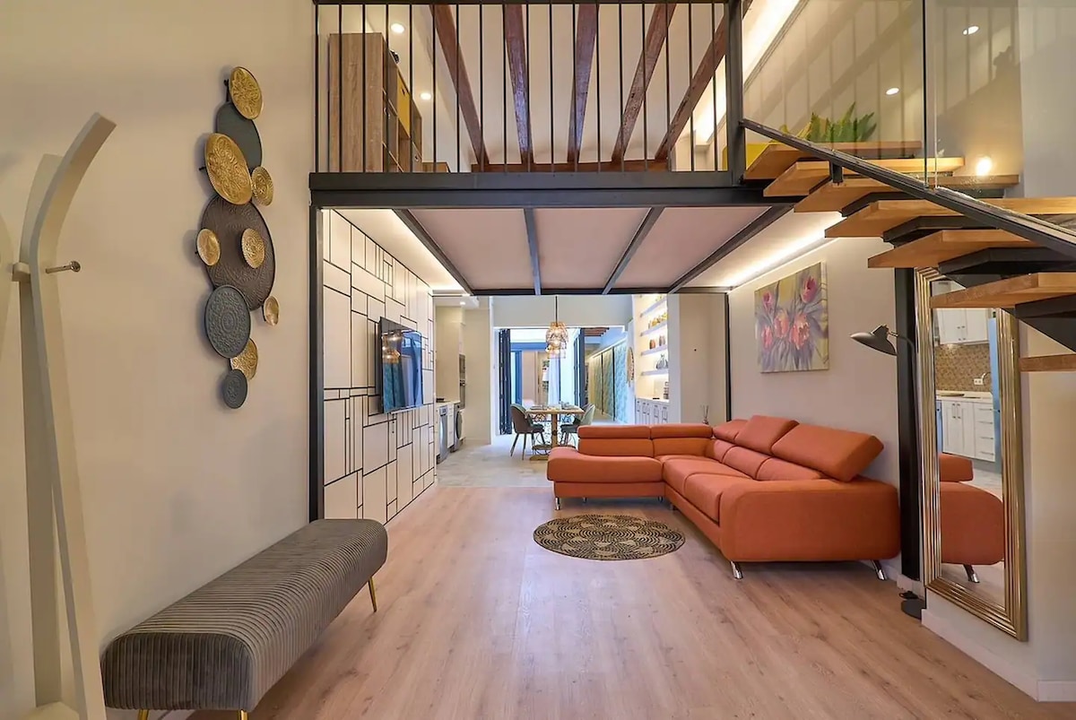 An open living area features a modern orange sectional sofa positioned around a circular rug. The area includes a wall-mounted TV and decorative wall art. A staircase leads to a lofted space, with additional seating and a dining area visible in the background.