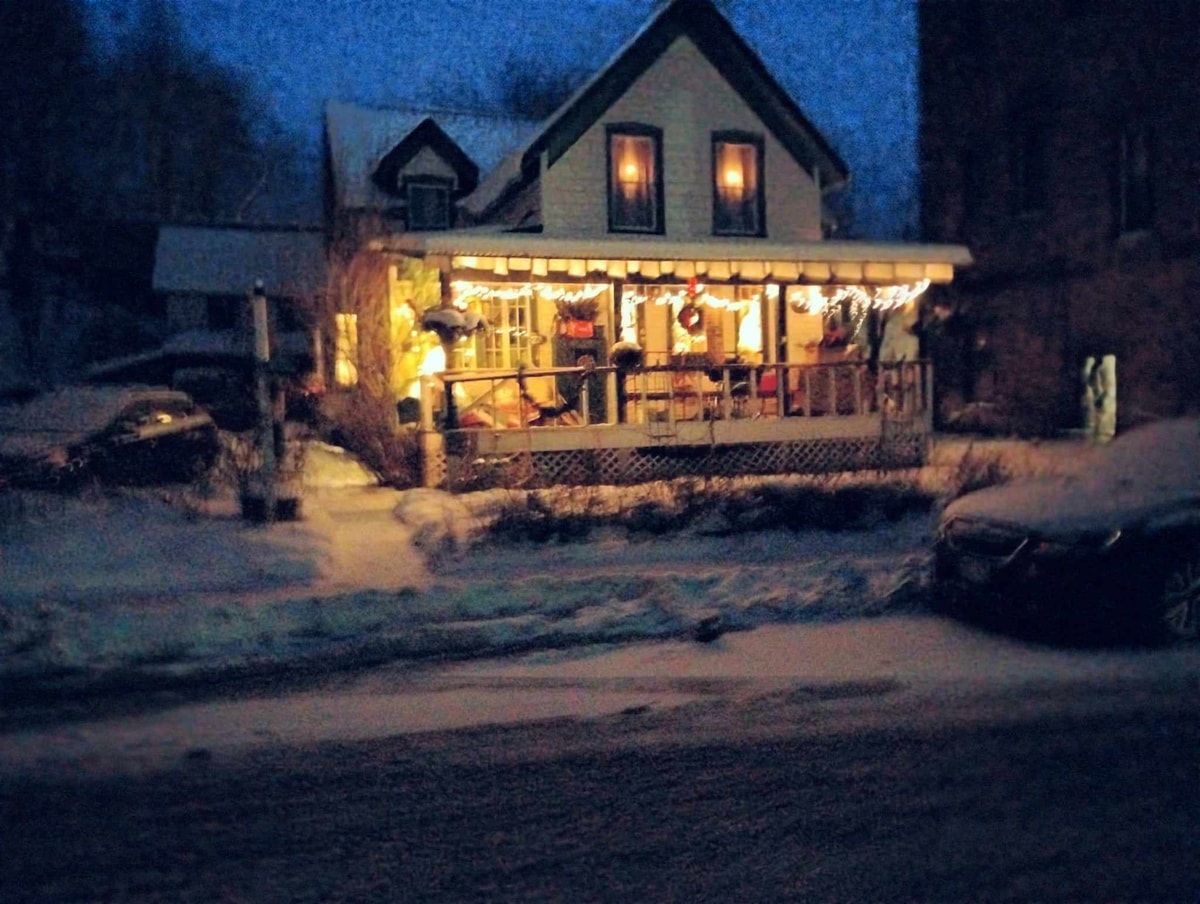 The exterior of a charming two-story house is illuminated softly in the evening light. A large front porch is adorned with festive decorations, while fresh snow blankets the ground, creating a serene winter scene. Nearby vehicles are parked along the snow-covered street.