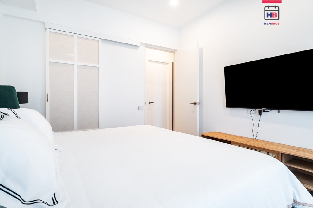 A minimalist bedroom is presented, featuring a spacious bed with crisp white linens. A large wall-mounted TV is positioned above a wooden media console. Soft, neutral tones create a calm ambiance, while a closed closet door and an open door leading to a hallway are visible.