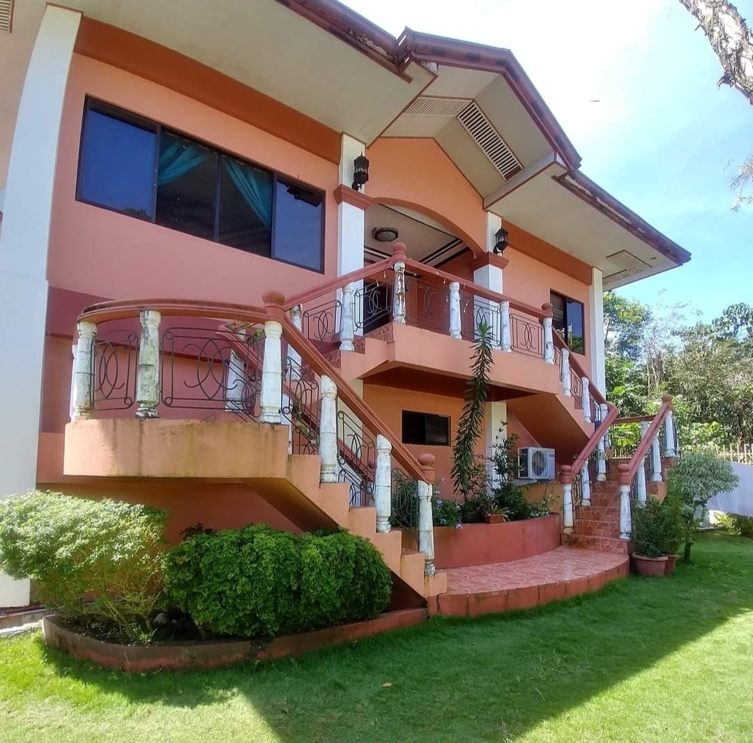 The exterior of a three-level home is shown, featuring a combination of soft pink and white walls. A landscaped grassy area surrounds the property, with elegant staircases leading to the balcony. Large windows and decorative railings enhance the inviting appearance of the home.