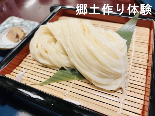 A serving of akita-style inaniwa udon is presented on a bamboo mat. The noodles are smooth and white, gently placed atop a decorative leaf. A small dish with dipping sauce is visible in the background, adding a local culinary touch.