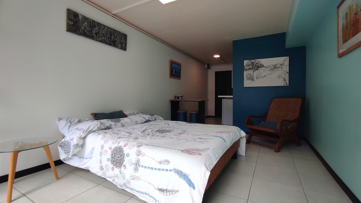 A spacious sleeping area features a double bed with a patterned duvet. An armchair is positioned nearby, and a small glass table sits beside the bed. The walls are painted in contrasting colors, creating a relaxed ambiance. A kitchenette is visible in the background.