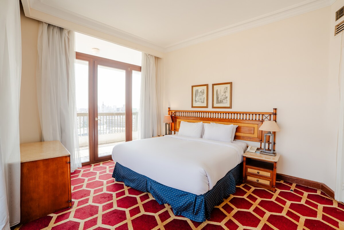 A spacious sleeping area features a large bed with crisp white linens, complemented by two bedside lamps. Natural light floods the room through double glass doors, which lead to a balcony. The floor is adorned with a geometric patterned carpet and a wooden dresser is positioned against a wall.