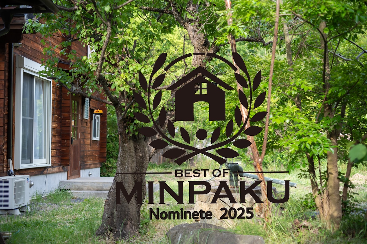 The exterior of the building features wooden paneling that harmonizes with the surrounding greenery. Large windows provide natural light, and lush trees frame the property, creating a serene outdoor atmosphere.