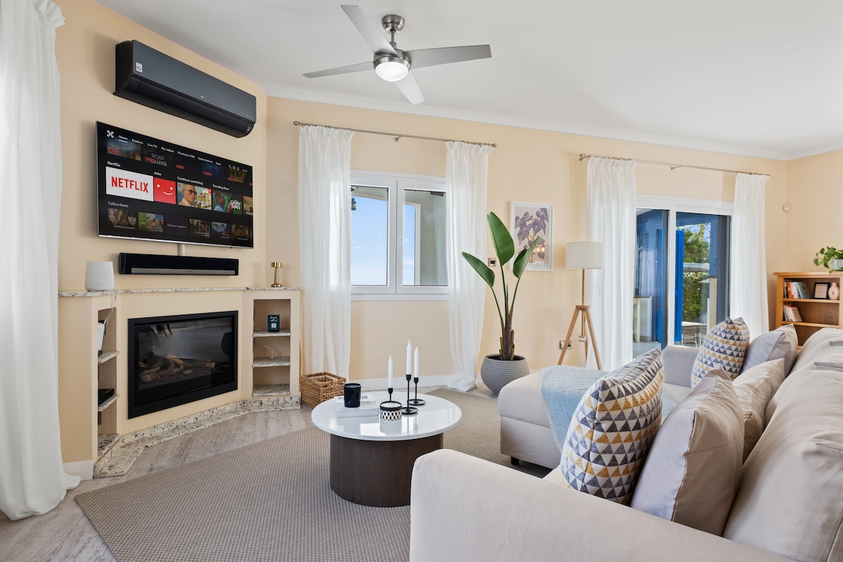 An open-plan living room features contemporary furnishings, with a large sectional sofa adorned with patterned pillows. A coffee table sits in the center, complemented by a wall-mounted TV. Bright, airy curtains frame the windows, allowing natural light to illuminate the space.