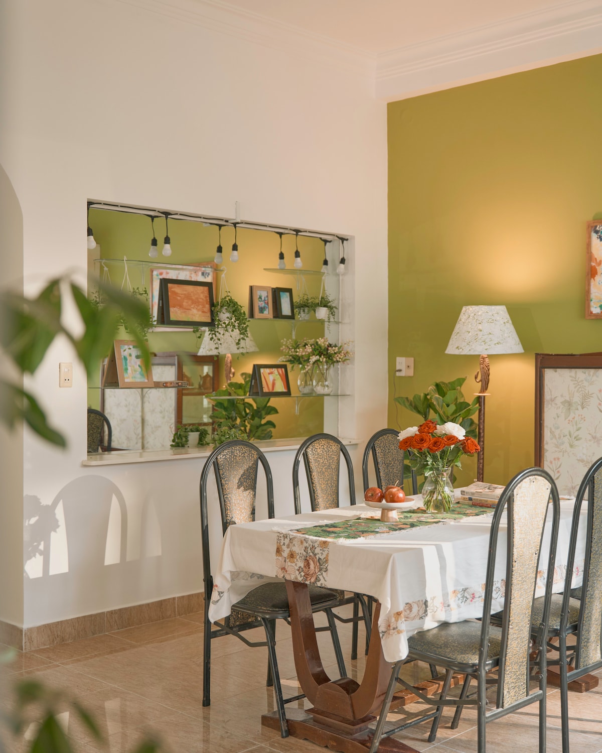A dining area is presented, featuring a rectangular table surrounded by six chairs. A tablecloth decorates the table, complemented by a vase of orange flowers. A wall display showcases framed artwork and plants, illuminated by soft lighting from a nearby lamp against a green wall.