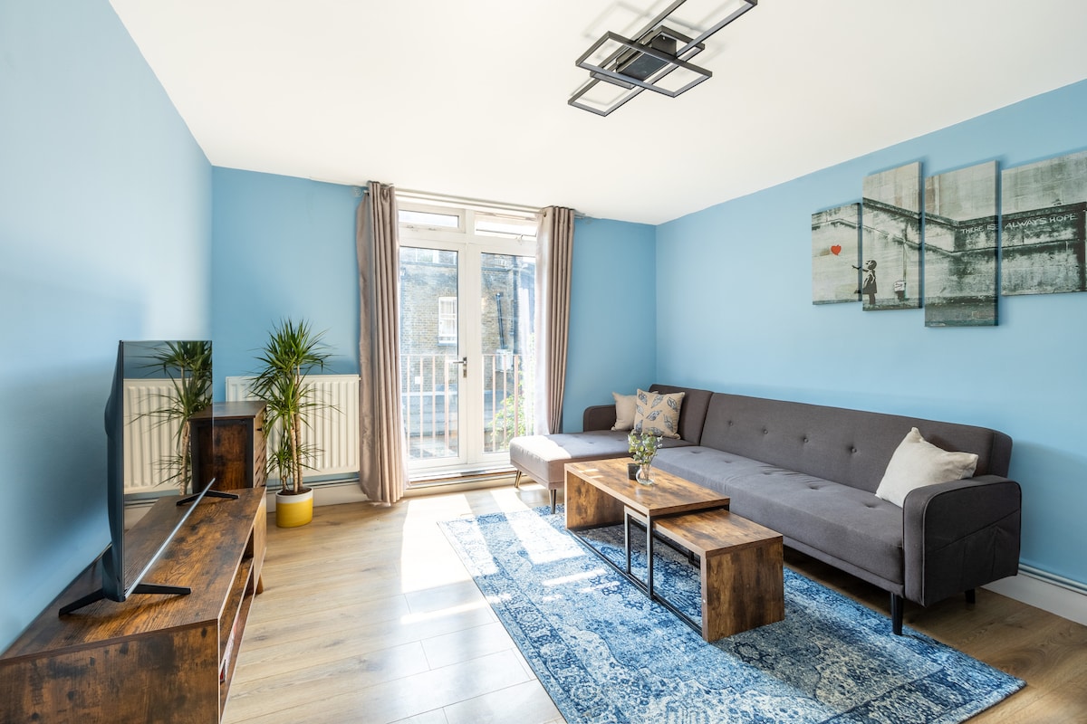 A spacious living area features light blue walls and large windows that allow natural light to fill the room. A gray sectional sofa is accompanied by a wooden coffee table, while a decorative area rug is placed beneath. Green plants add a touch of nature to the space.