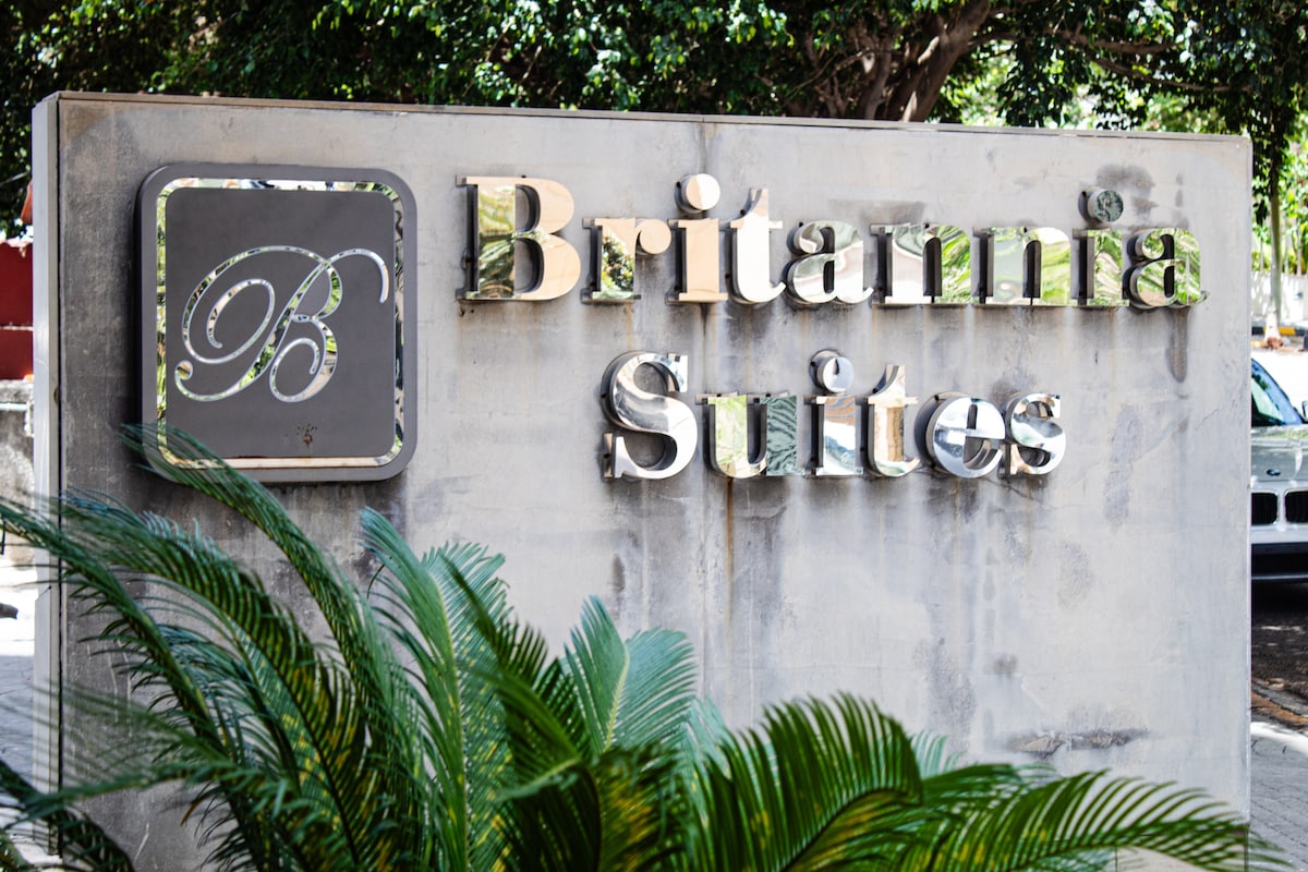 The entrance sign of Britannia Suites is prominently displayed, featuring elegant lettering against a textured backdrop. Lush green foliage is visible at the base, complementing the modern design of the sign and creating an inviting welcome.