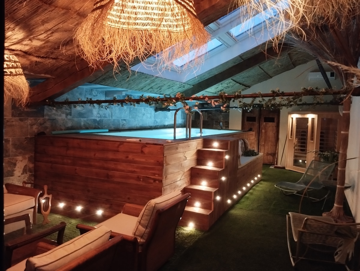 An indoor pool is featured within a rustic space with exposed wooden beams and stone walls. Soft lighting highlights the wooden structure surrounding the pool, while green accents of artificial grass and comfortable seating create an inviting area for relaxation.