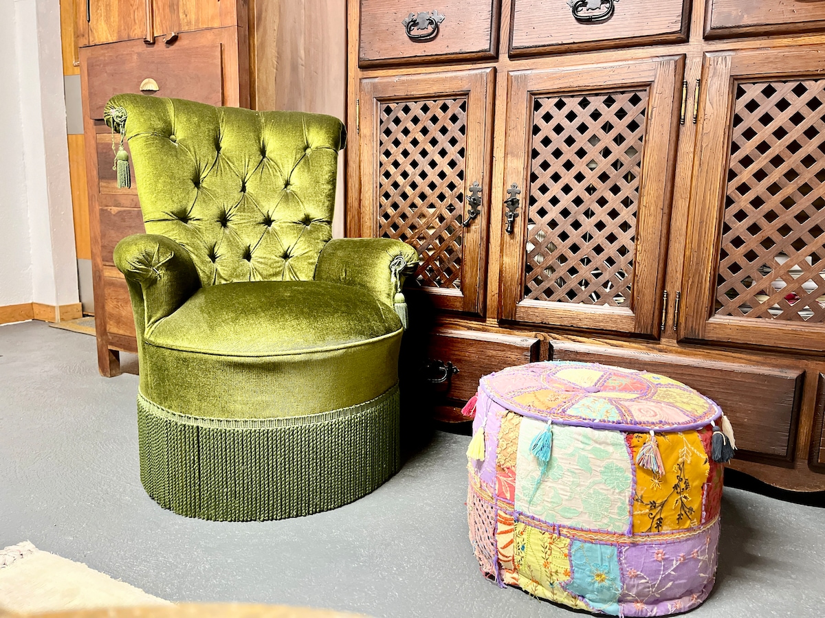 A vintage green armchair with tufted upholstery is positioned beside a colorful, round pouf featuring intricate patterns and tassels. Behind the furniture, a wooden cabinet with detailed latticework provides additional texture and warmth to the space.