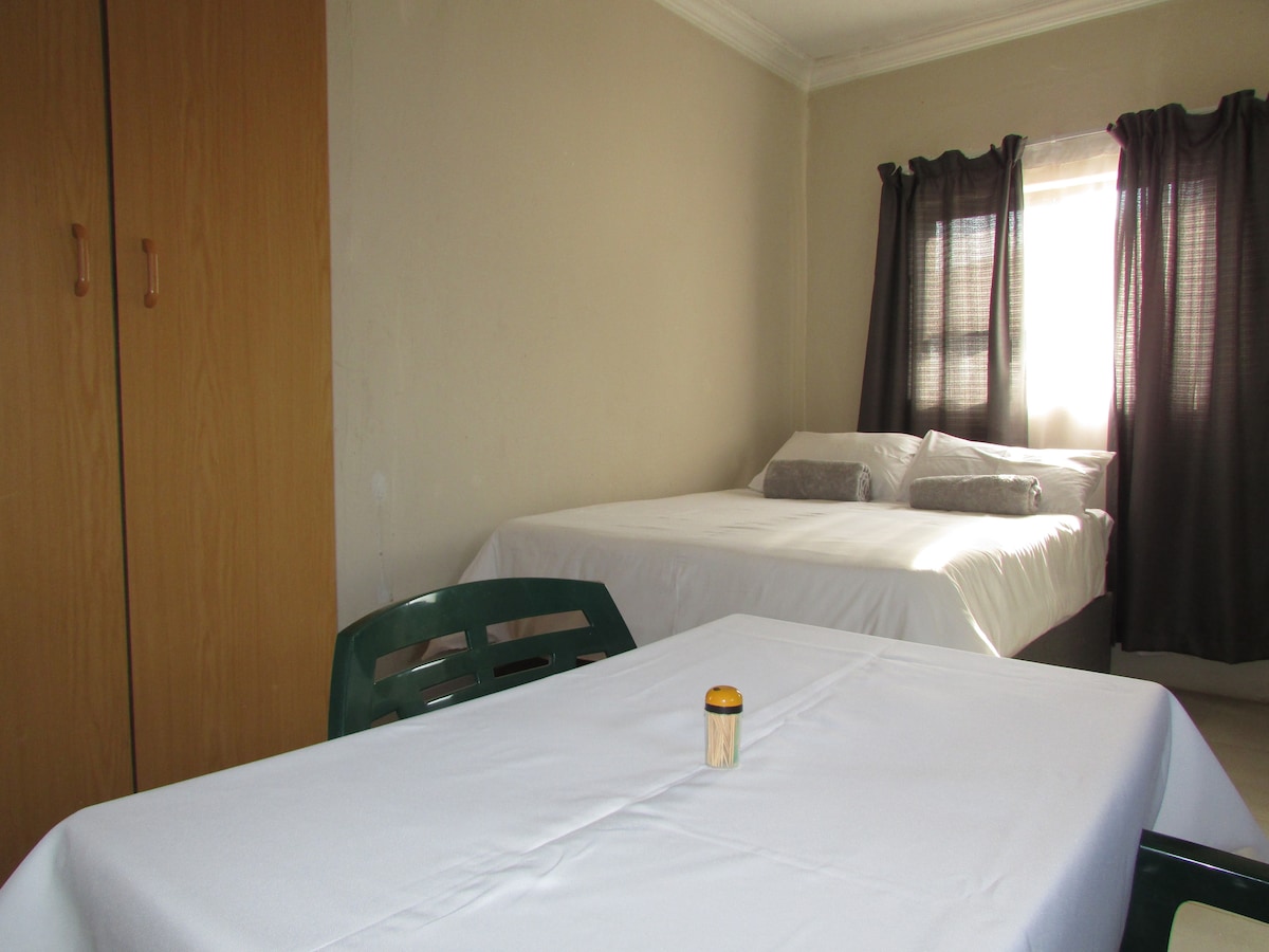 A simple and functional sleeping area features a spacious bed with light bedding and two accent pillows. A neatly arranged dining table with a white tablecloth sits in the foreground. Two wooden wardrobes are positioned against the wall, and natural light enters through a partially curtained window.
