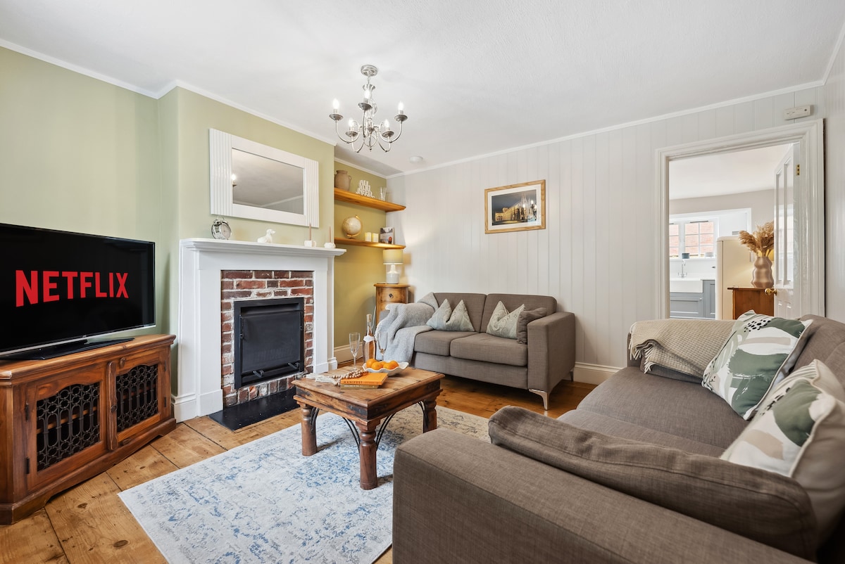 The living area features two comfortable sofas and a wooden coffee table. A large television displays Netflix, and a decorative fireplace is complemented by a brick wall. Natural light enters through the doorway leading to the rest of the home, enhancing the warm ambiance.