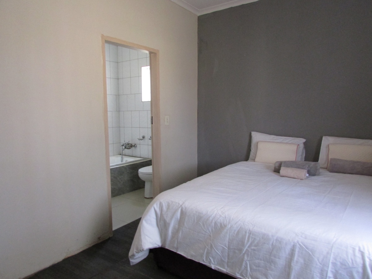 A spacious sleeping area features a neatly made bed with light bedding and a pair of plush pillows. A door leads to an adjoining bathroom, which includes a bathtub and a toilet, visible through the open entryway.