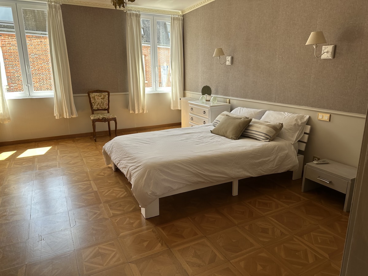 A spacious bedroom is illuminated by natural light from large windows, featuring light-colored walls and warm wooden flooring. A simple bed is centered in the room, accompanied by bedside tables and a decorative armchair. Two wall-mounted lamps provide soft lighting.