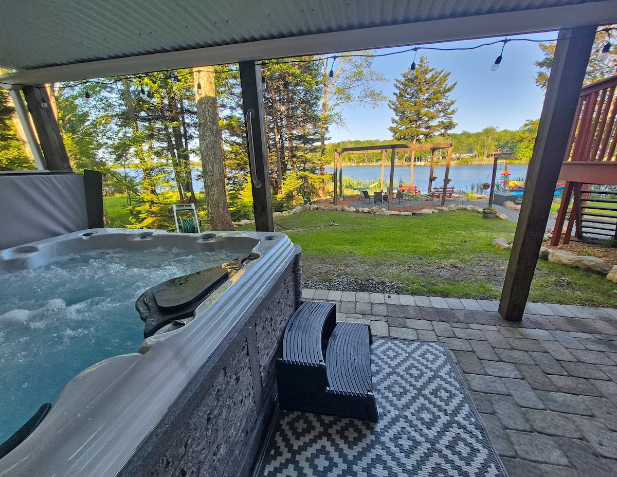 An outdoor hot tub sits on a paved surface, with steps leading up to it. Beyond, a scenic view of the lake is visible, framed by tall trees. A shaded area features a gathering spot with colorful chairs and a pergola, providing a serene outdoor retreat.