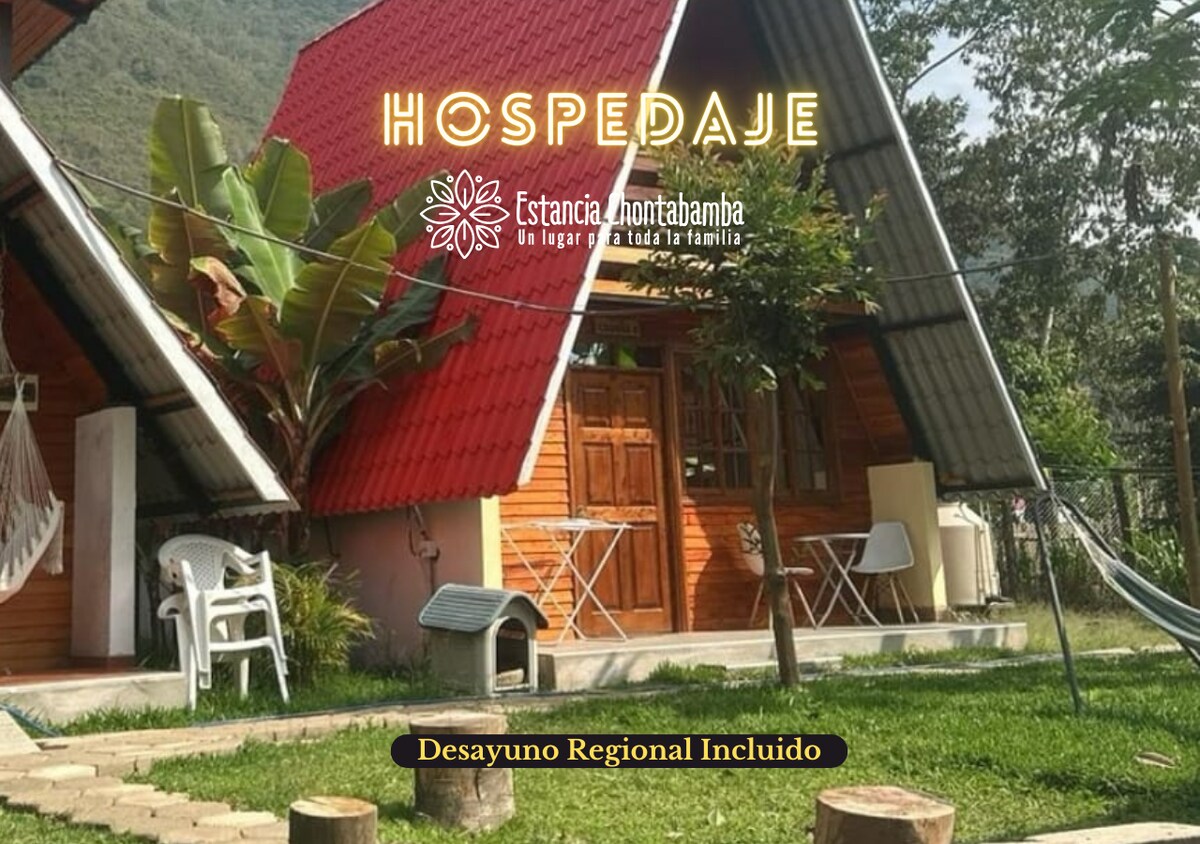 A charming A-frame cabin with a red roof is surrounded by greenery. A well-maintained lawn features tropical plants and a hammock, offering a relaxed outdoor space. The overall design reflects a cozy, family-friendly environment, inviting tranquility amidst the natural landscape.