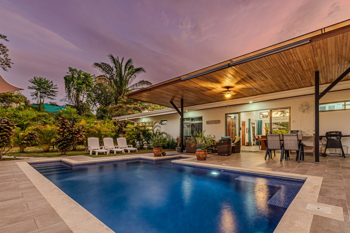 A private outdoor area features a tranquil blue pool surrounded by lounge chairs under a wooden-covered patio. The space is complemented by lush greenery in the background, providing a serene atmosphere for relaxation.
