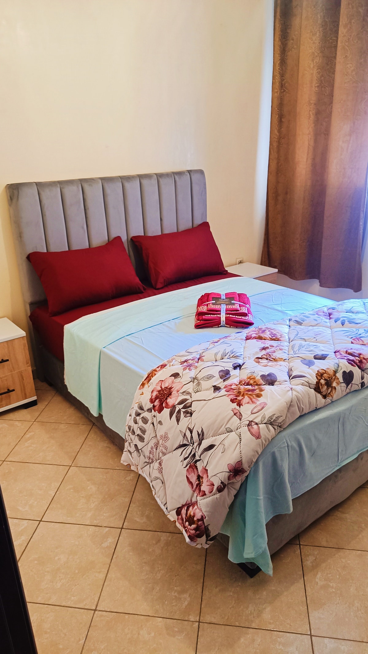 A cozy bedroom features a spacious bed with a soft, grey headboard, adorned with red pillows. A colorful floral quilt covers the bed, complemented by light blue bedding. A nightstand is positioned nearby, and sunlight filters through a curtain, illuminating the warm space.