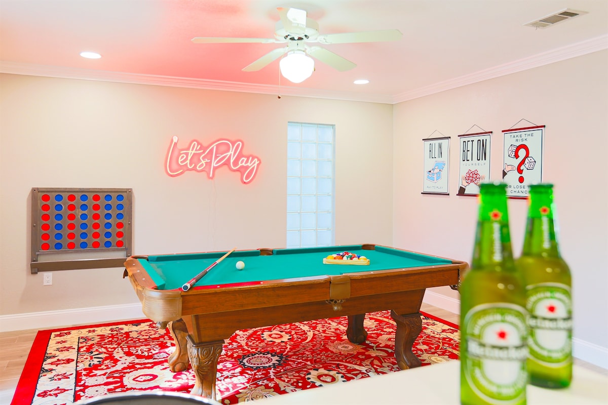 A game room features a classic pool table centered on a patterned area rug. A 'Let's Play' neon sign adds a playful touch to the space, complemented by wall art. A connect-four game is positioned against the wall, enhancing the enjoyable atmosphere.