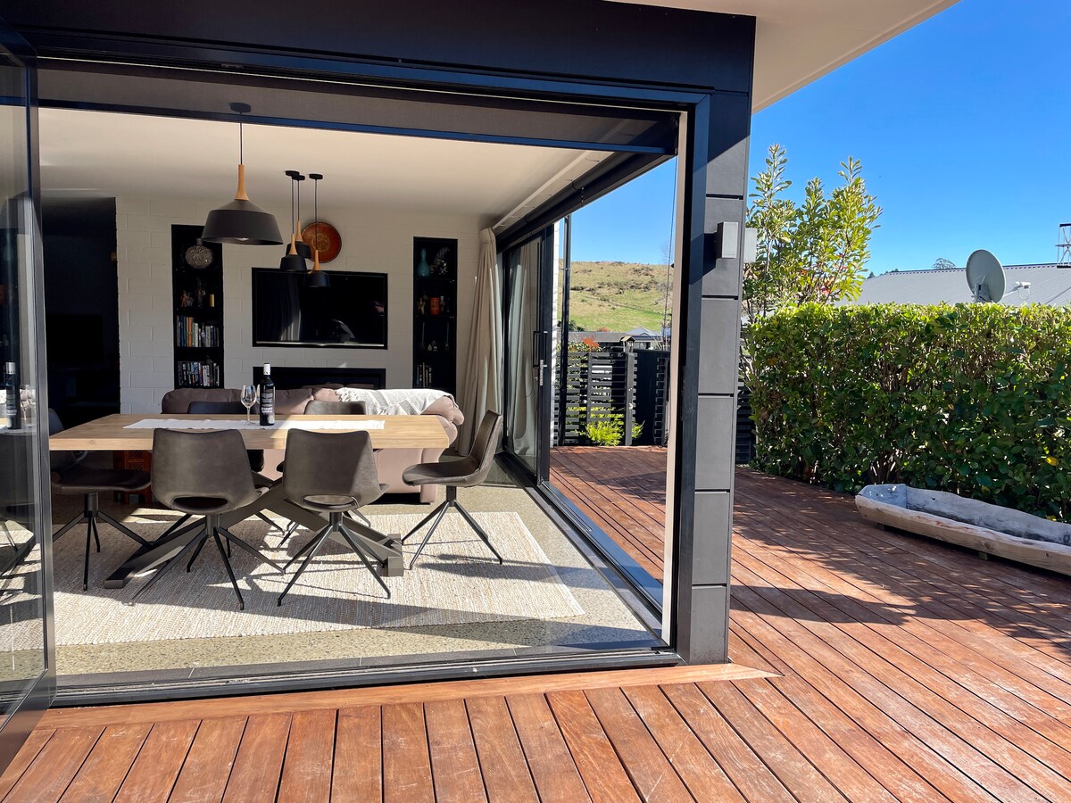 A dining area with a large wooden table and comfortable chairs is viewed through expansive glass doors. Natural light floods the space, which includes a TV mounted on the wall. A wooden deck extends outside, surrounded by lush greenery.