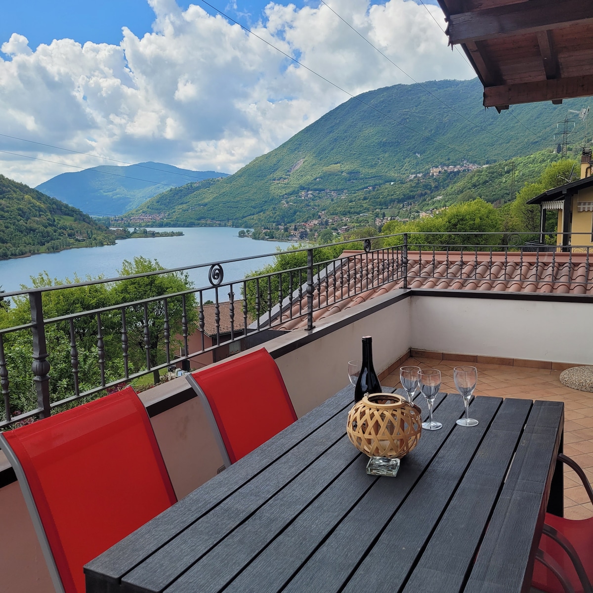 A spacious terrace overlooks Lake Endine, featuring a large table with six red chairs. A centerpiece made of woven material sits atop the table, accompanied by glasses and a bottle. Lush green hills frame the tranquil lake under a partly cloudy sky.