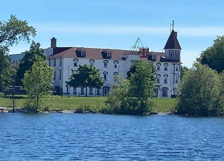 A historic building is viewed from across a body of water, surrounded by lush green trees. The structure features a classic architecture with a distinctive tower, multiple windows, and a sloped roof, set against a clear blue sky.