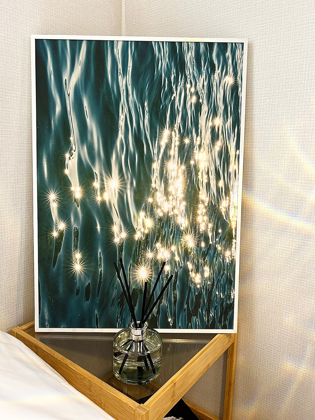 A framed artwork depicting shimmering water reflections is displayed on a wall. A glass vase with reed diffusers sits on a wooden nightstand beside a spacious bed, creating a soothing atmosphere.
