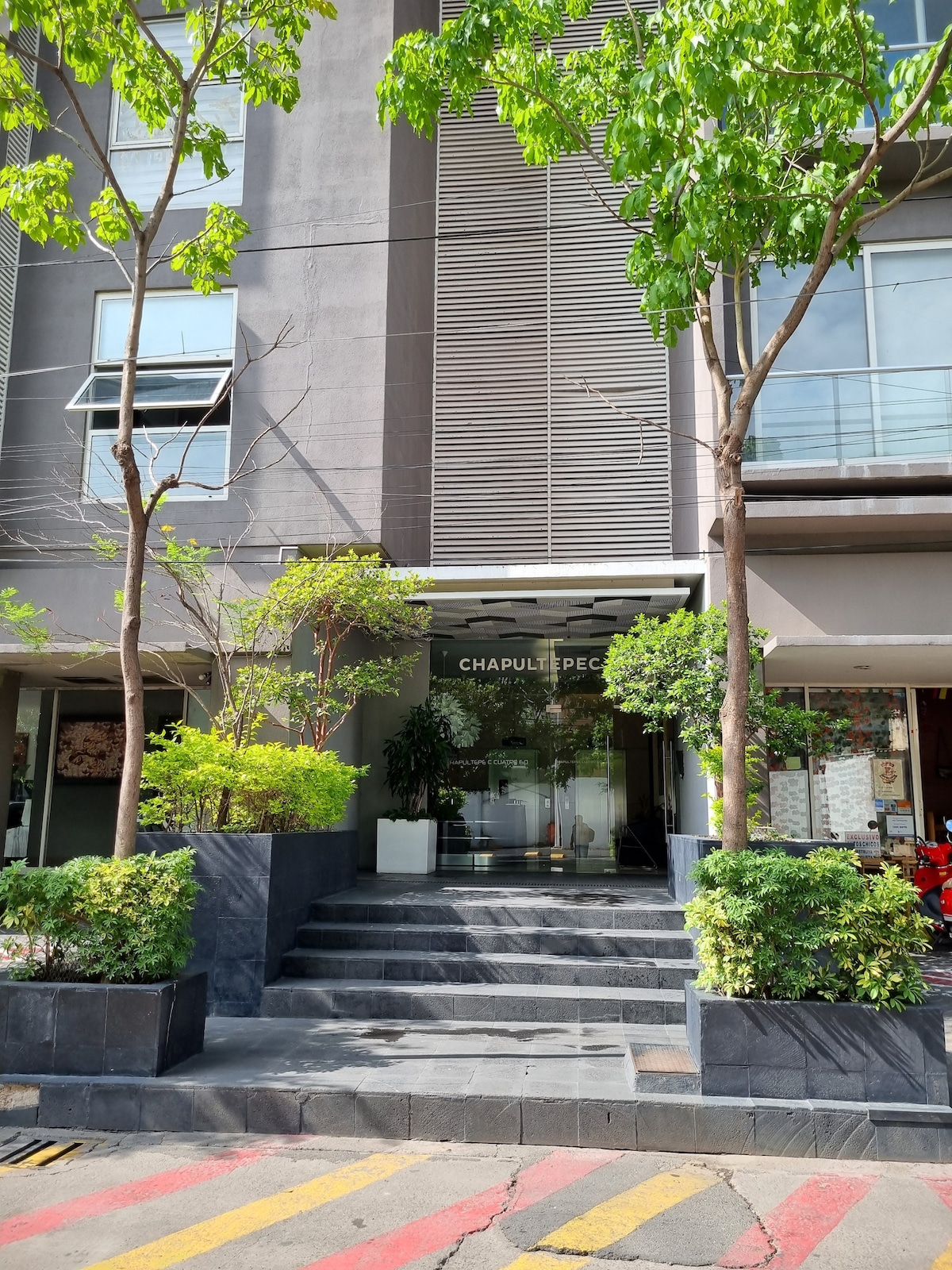 The entrance to the modern residential tower is framed by green trees and neatly landscaped plants. A set of wide steps leads to a glass door marked with the name 'Chapultepec.' The building's façade features a mix of textured panels and large windows.