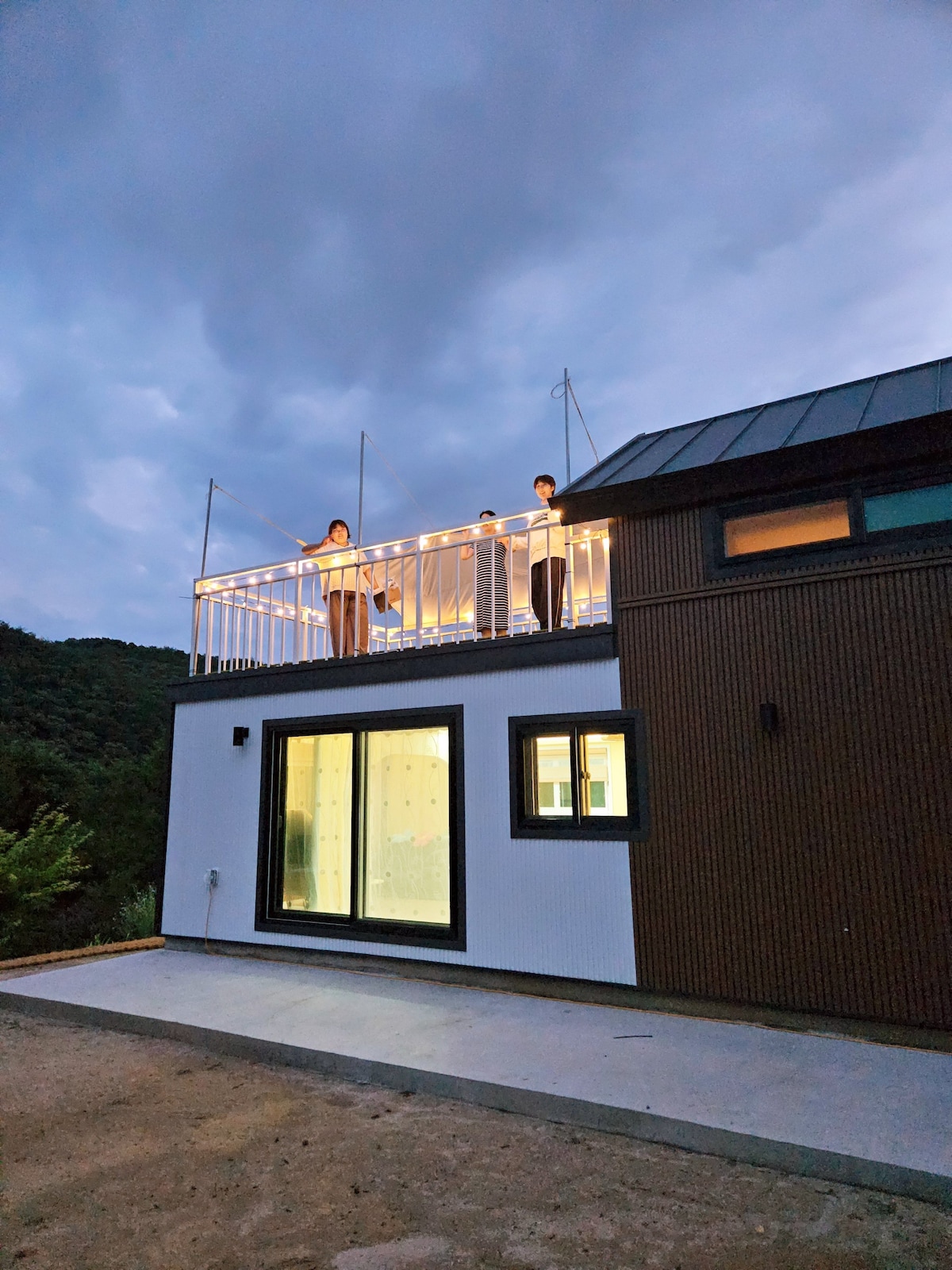 A modern two-story structure features a spacious rooftop deck adorned with soft lighting, offering scenic views of the surrounding nature. Large windows illuminate the interior, with the contrasting wooden and white facade enhancing the building's contemporary design.