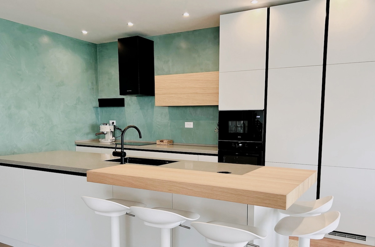 A modern kitchen features a central island with a wooden countertop, complemented by white bar stools. Sleek cabinetry in muted tones contrasts with the textured green wall, while built-in appliances and a minimalist design create an inviting cooking space.