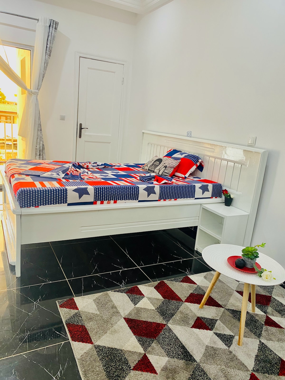 A cozy bedroom features a spacious bed with a colorful patterned duvet cover and decorative pillows. A small bedside table holds a lamp and decorative items. Natural light enters through a nearby window, and a patterned rug adds a touch of warmth to the dark floor.