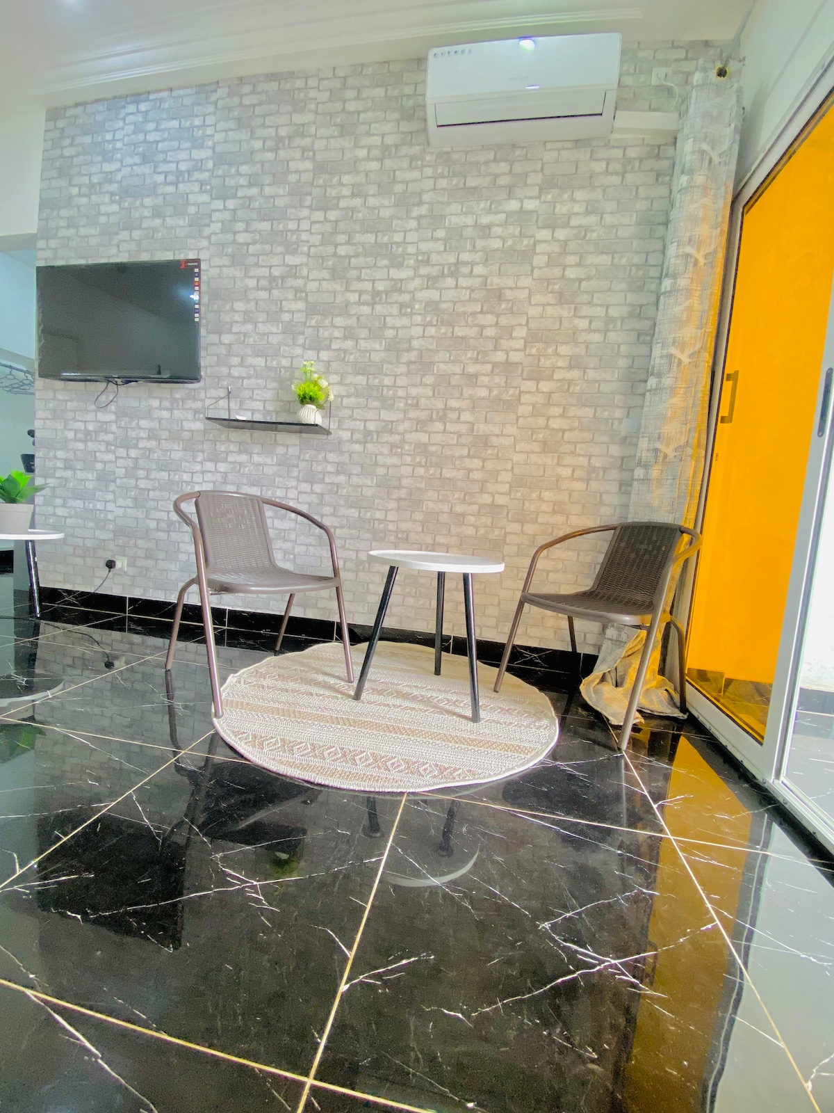 A cozy sitting area is presented with two woven chairs set around a small round table. The dark tiled floor reflects the modern decor, while a textured gray wall adds visual interest. A flat-screen TV is mounted nearby, and natural light filters in through a glass door.