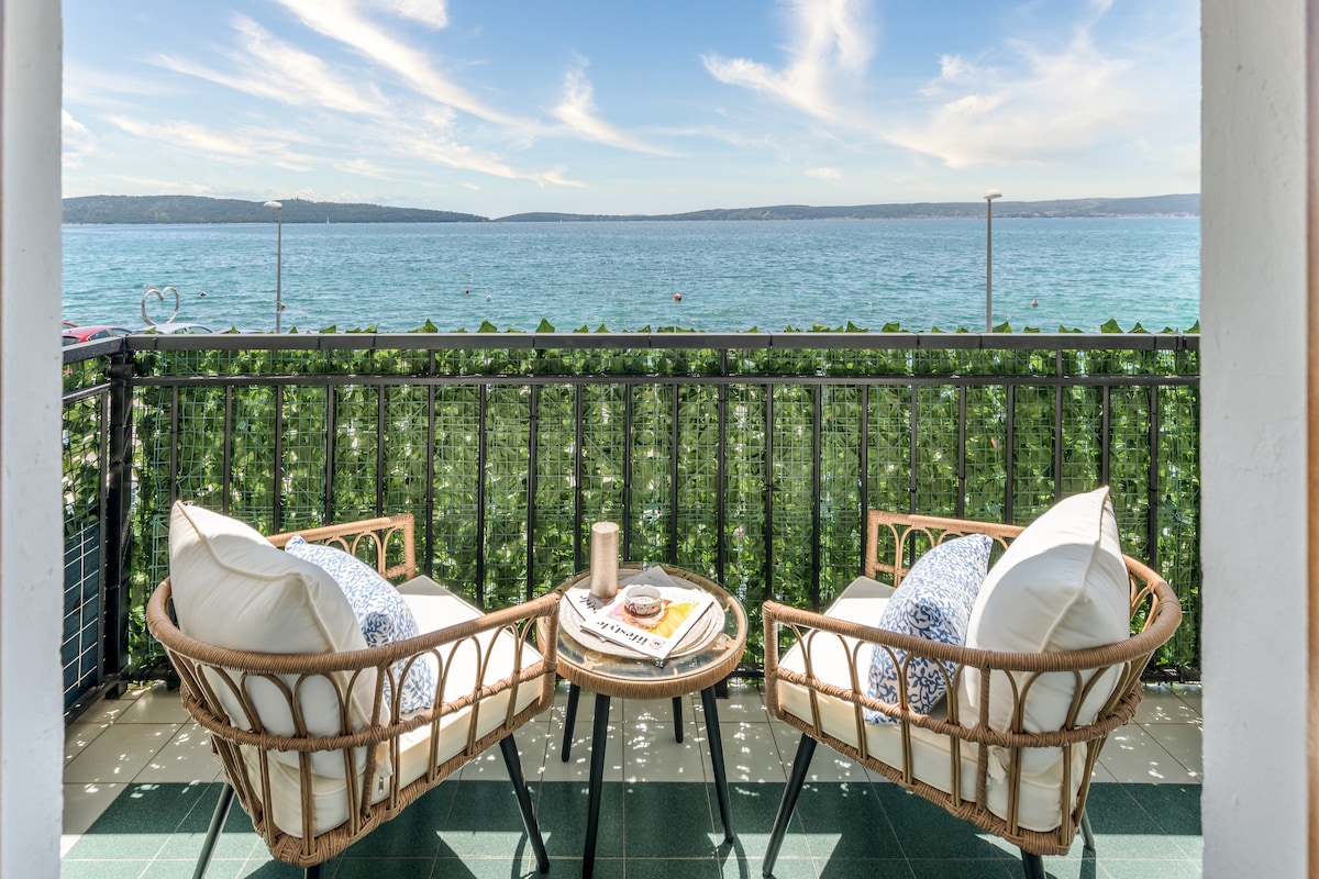 A south-facing balcony provides unobstructed sea views, featuring a round table surrounded by two wicker chairs. Blue cushions are placed on each chair, with a magazine and glass on the table, inviting relaxation while enjoying the coastal scenery.