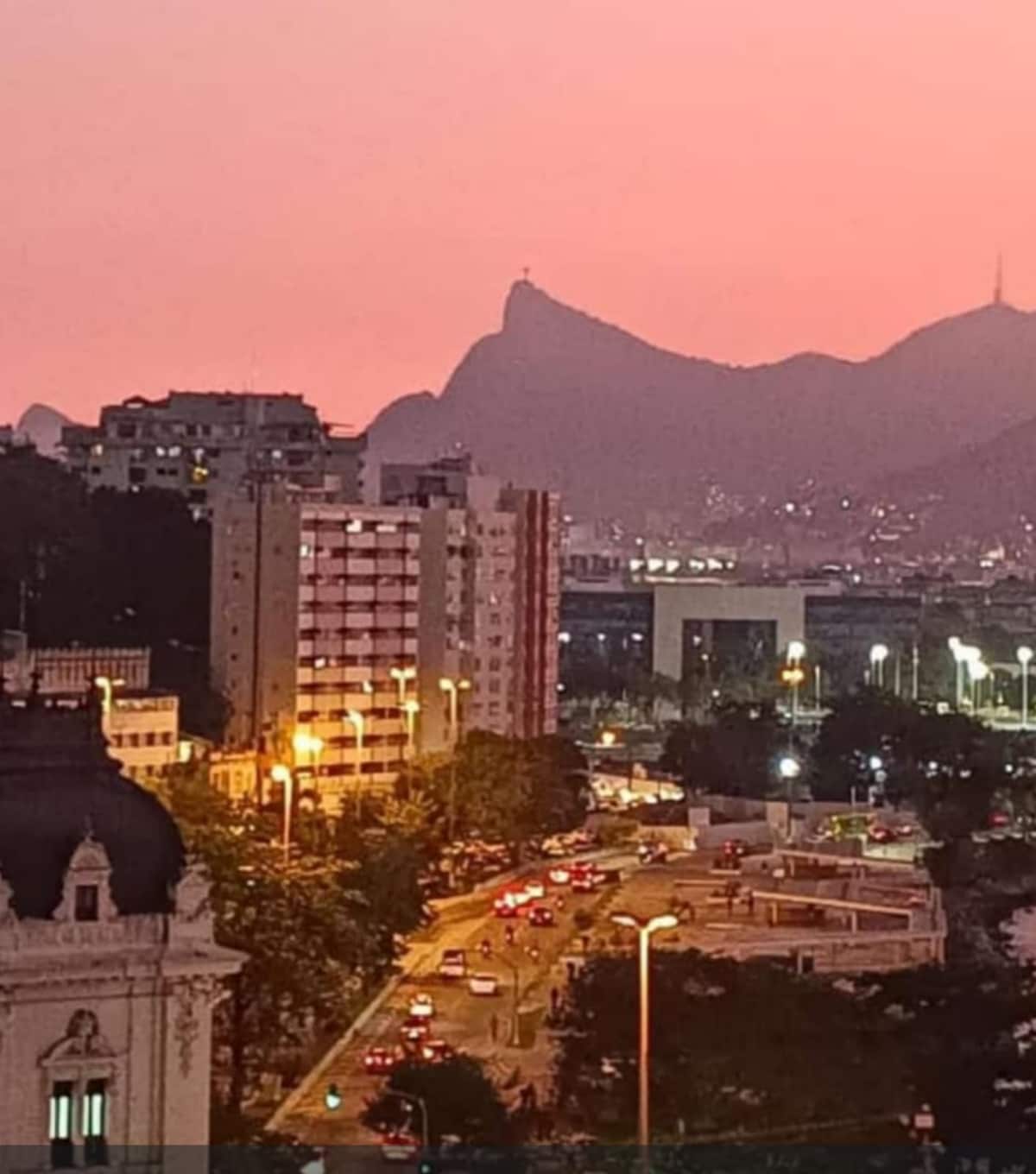 A breathtaking evening view captures a cityscape at sunset, featuring a blend of urban buildings and natural mountains. Warm orange and pink hues fill the sky, while city lights begin to illuminate the streets below, creating a serene atmosphere of Niterói.