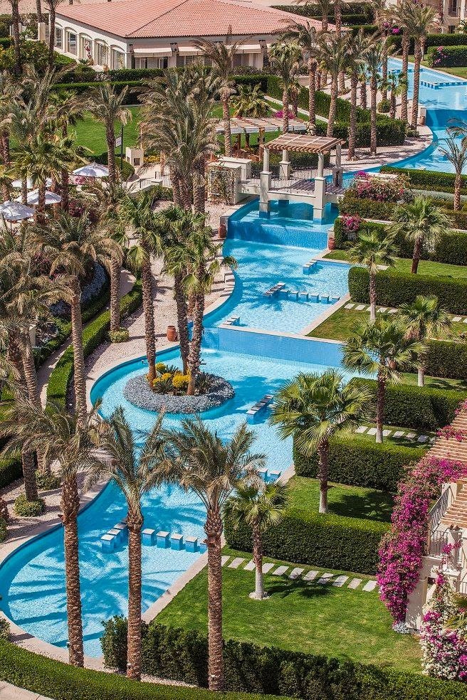 A serene outdoor space features winding turquoise pools bordered by manicured landscaping and palm trees. Vibrant bougainvillea adds color to the area, which includes lounging chairs placed beside the water. This garden-like setting is designed for relaxation amidst a tranquil environment.