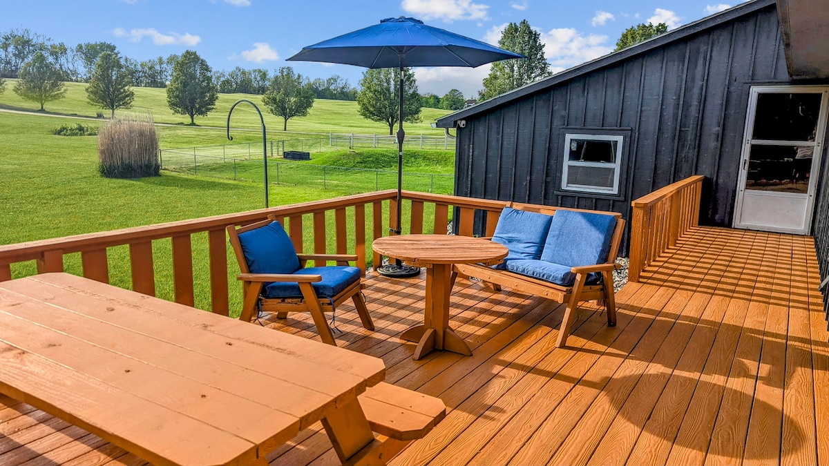 The spacious outdoor deck features wooden flooring with multiple seating areas, including a round table and two lounge chairs. An umbrella provides shade, while green grassy fields and trees are visible in the background, creating a connection to nature.