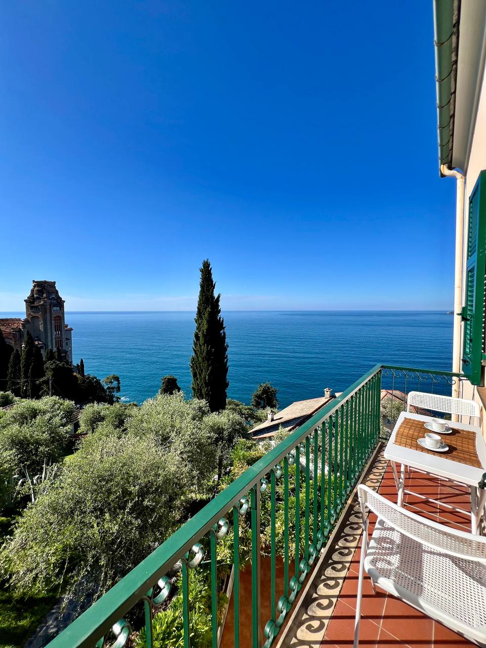 A balcony with green railings overlooks the serene sea, dotted with a few distant boats. A table is set with two chairs, inviting relaxation amid olive trees and lush greenery. The clear blue sky complements the tranquil coastal view, enhancing the peaceful ambiance.
