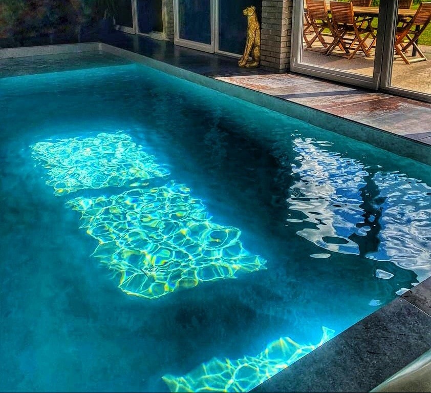 A spacious indoor pool is visible, featuring shimmering water that reflects light patterns on the surface. Large glass doors open to an outdoor area, where wooden lounge chairs can be seen nearby against a backdrop of greenery.