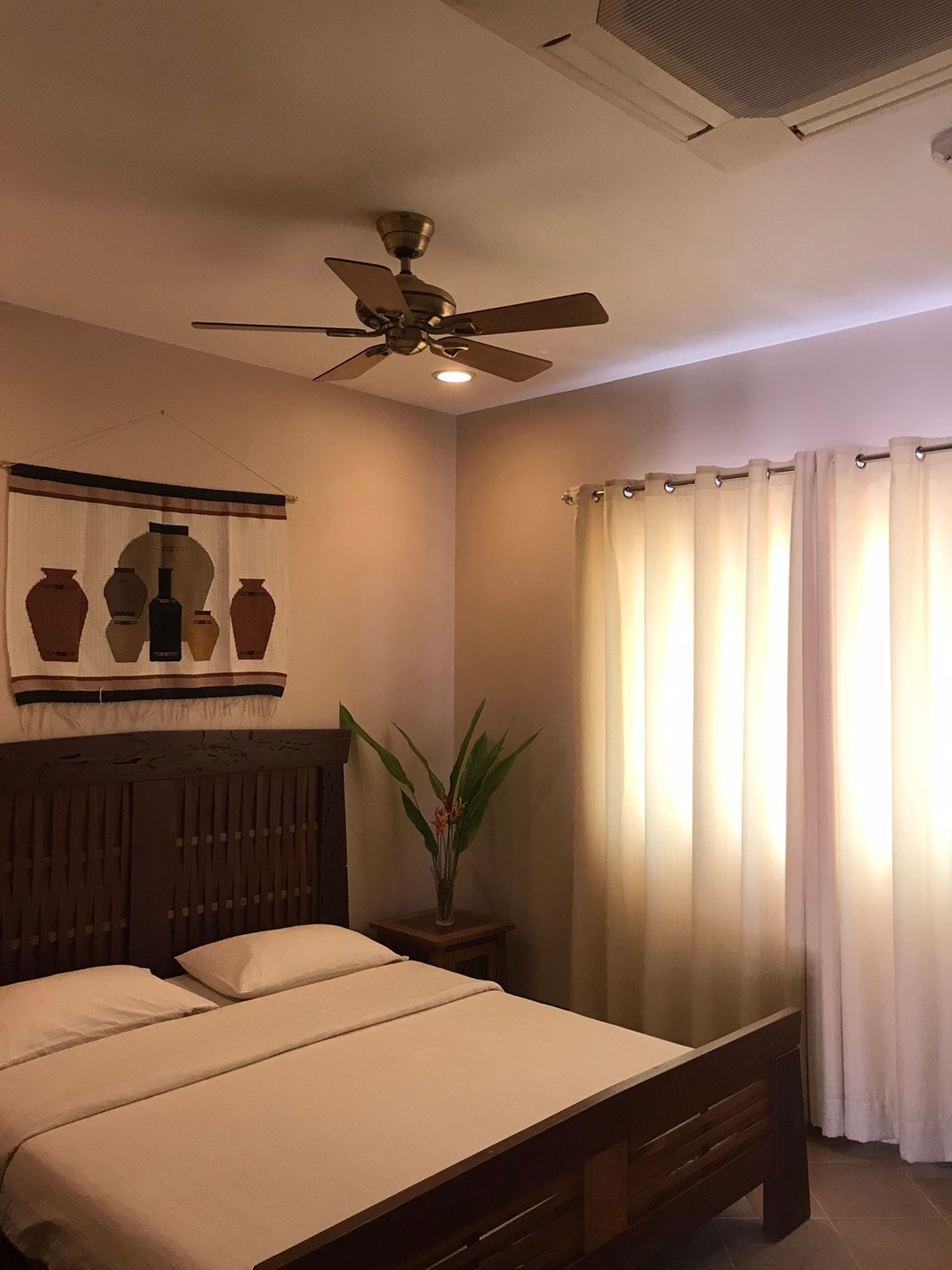 A serene bedroom is featured with a wooden bed framed by neutral-colored walls. A ceiling fan is visible above, and natural light streams through sheer curtains. Decorative wall art with vases adds an artistic touch, while a small plant sits beside the bed.