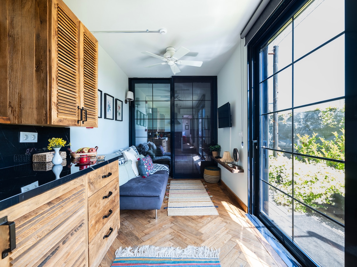 A cozy seating area is featured, enhanced by natural light from large glass doors. Wooden cabinetry holds decorative items, and a soft fabric sofa invites relaxation. Artwork is displayed on the walls, complementing the warm tones of the wooden floor with a woven rug adding texture.