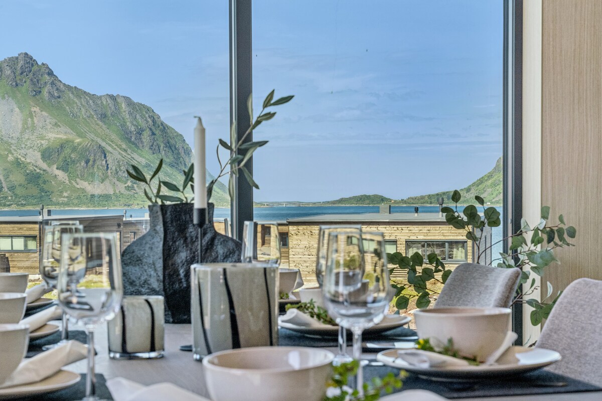 A dining area is presented with elegantly arranged table settings, featuring white plates and glassware. Behind, expansive windows frame a stunning view of lush mountains and the coastline, inviting natural light into the space.