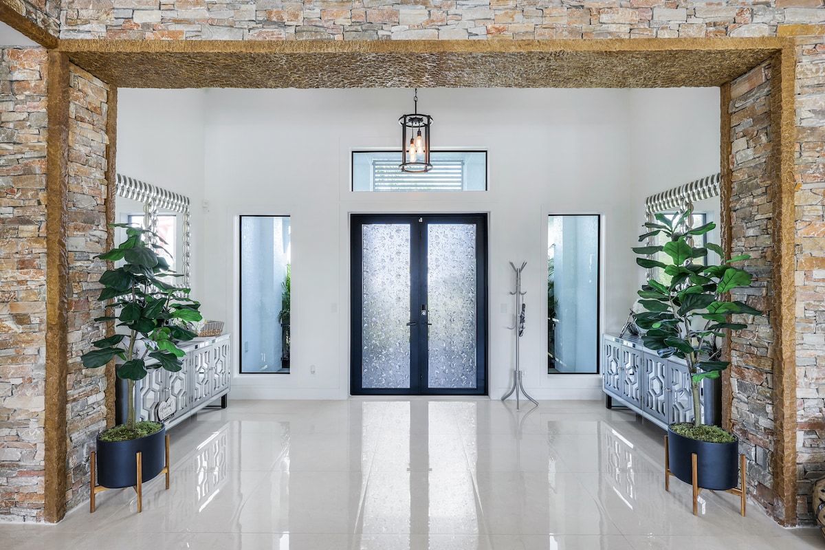 An expansive entryway is framed by natural stone walls. Two large mirrors reflect light, while stylish cabinetry with intricate designs is placed on either side. Lush potted plants add a touch of greenery, complemented by polished floor tiles that enhance the spacious ambiance.