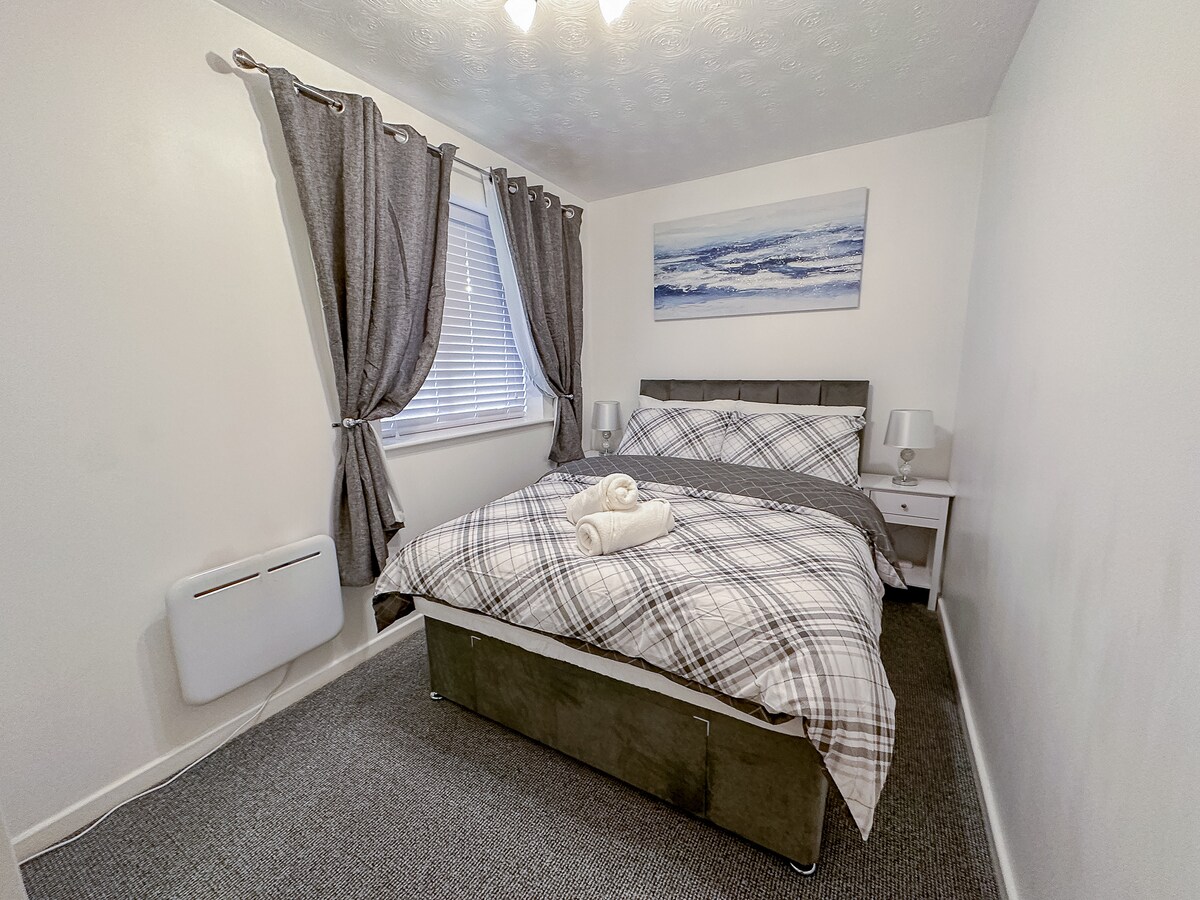 A well-lit bedroom features a spacious bed dressed in a neutral plaid blanket, with a folded towel placed neatly on top. Two bedside tables are positioned beside the bed, each topped with a lamp. Dark curtains frame the window, allowing natural light to filter in.