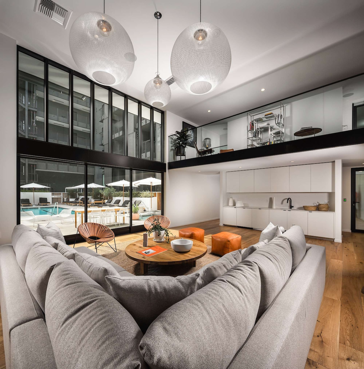 A spacious living area features large glass windows that offer views of the pool area. The room is furnished with a comfortable sectional sofa and a wooden coffee table, complemented by decorative lighting and an open kitchen space, enhancing the modern design.