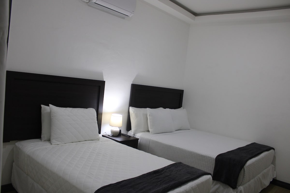 A well-lit room features two comfortable beds with crisp white linens. A bedside table with a lamp is positioned between the beds. The walls are painted in a neutral tone, and a ceiling air conditioning unit is present.