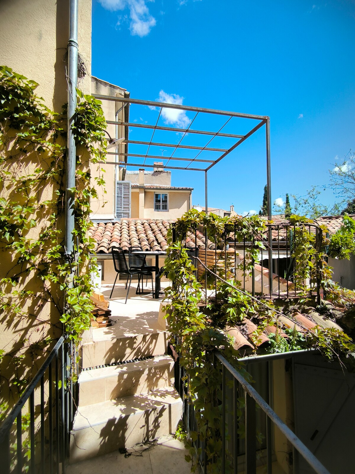 A terrace with a metal frame overlooks the surrounding rooftops, bordered by lush green vines. A small dining table with four chairs is situated on the terrace, providing an inviting space for outdoor meals. The clear blue sky adds to the relaxing ambiance.