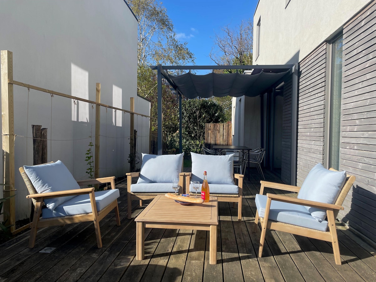 A welcoming outdoor seating area is depicted, featuring four soft blue chairs paired with a wooden coffee table. A shade structure covers part of the space, enhancing comfort. The surroundings include greenery, providing a serene atmosphere ideal for relaxation.